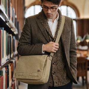 Man in a library wearing a tweed blazer and a unisex khaki canvas satchel with a minimalist black square logo.