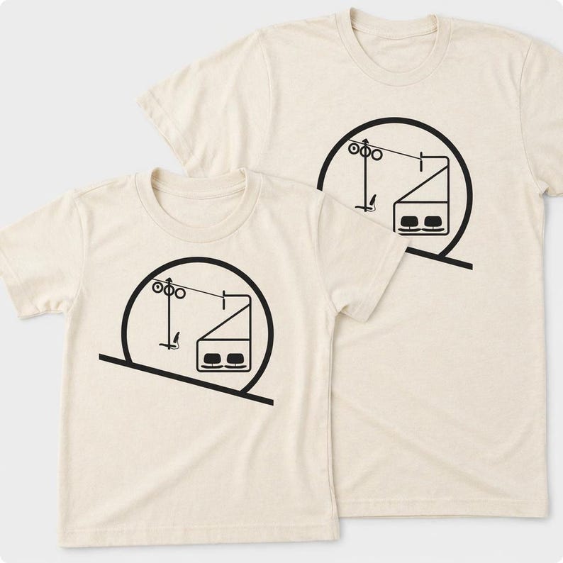 May include: Two cream-colored short-sleeve t-shirts featuring a black graphic print of a ski lift chair within a circular frame. The design depicts a cable car system with a single seat and a larger cabin, set against a plain, light-colored fabric.