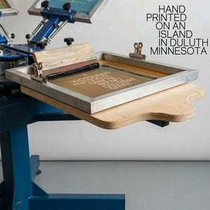 May include: A blue screen printing press with a metal frame and a wooden squeegee. The screen features the text HAND PRINTED ON AN ISLAND IN DULUTH MINNESOTA. The equipment includes metal clamps and a wooden platen for holding the printing surface.