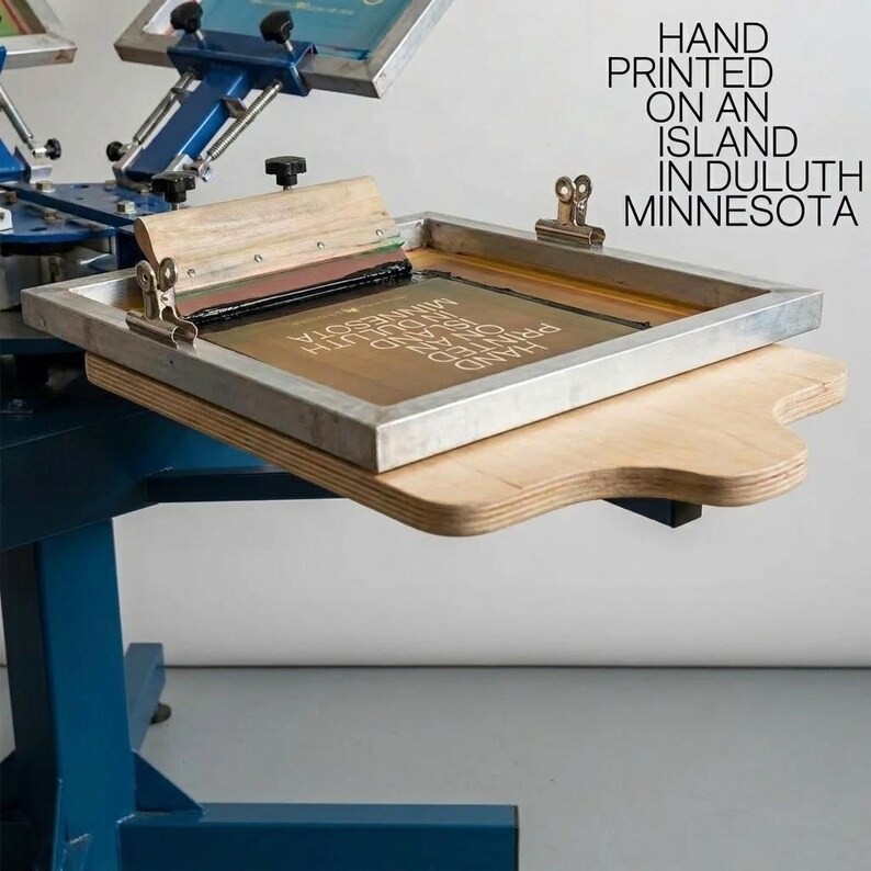 May include: A blue screen printing press with a metal frame and a wooden squeegee. The screen features the text HAND PRINTED ON AN ISLAND IN DULUTH MINNESOTA. The equipment includes metal clamps and a wooden platen for holding the printing surface.