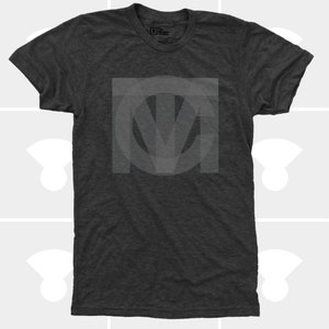 A flat-lay product photo of a premium dark charcoal grey tri-blend t-shirt featuring a large, subtle tonal interlocking geometric TMC monogram logo screen-printed on the center chest.