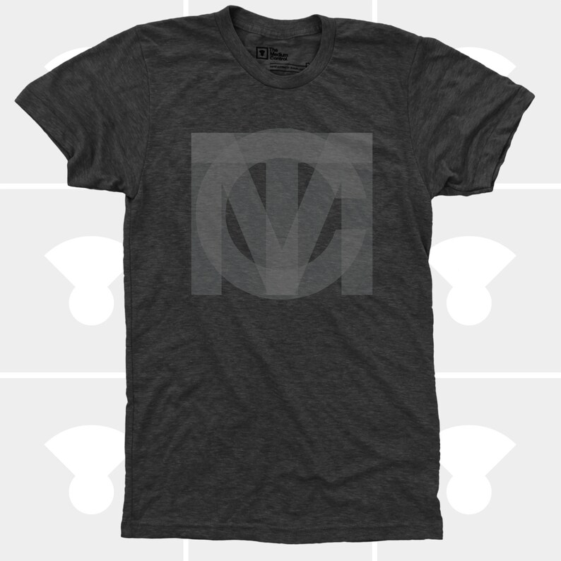 A flat-lay product photo of a premium dark charcoal grey tri-blend t-shirt featuring a large, subtle tonal interlocking geometric TMC monogram logo screen-printed on the center chest.