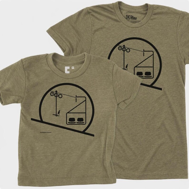May include: Two olive green heathered t-shirts featuring a black graphic print of a ski lift chair within a circular frame. The design includes a support tower, cables, and a two-seat chair. The text "The Medium Control" is printed on the shirts.