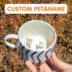 Peut inclure: Une tasse en céramique blanche avec un motif ondulé bleu et blanc. À l'intérieur de la tasse se trouvent deux petites figurines en céramique de chats. La tasse est tenue par une main.