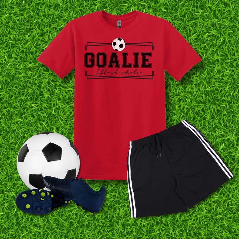 Puede incluir: Camiseta roja con un gr&aacute;fico en blanco y negro que dice "Goalie I block shots" y un gr&aacute;fico de bal&oacute;n de f&uacute;tbol. Un pantal&oacute;n corto deportivo negro con rayas blancas a los lados. Un bal&oacute;n de f&uacute;tbol blanco y negro y un par de botas de f&uacute;tbol azules.