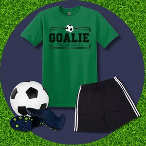 Puede incluir: Camiseta verde con un gr&aacute;fico de bal&oacute;n de f&uacute;tbol blanco y negro y el texto "GOALIE I block shots". Tambi&eacute;n se muestra un bal&oacute;n de f&uacute;tbol negro, tacos de f&uacute;tbol azules y pantalones cortos deportivos negros con rayas blancas.