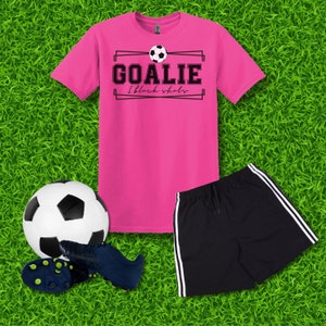 Puede incluir: Camiseta rosa con texto negro que dice "Goalie I block shots" y un gr&aacute;fico de bal&oacute;n de f&uacute;tbol blanco y negro. Un pantal&oacute;n corto deportivo negro con rayas blancas a los lados. Un bal&oacute;n de f&uacute;tbol blanco y negro y un par de tacos de f&uacute;tbol negros y azules.