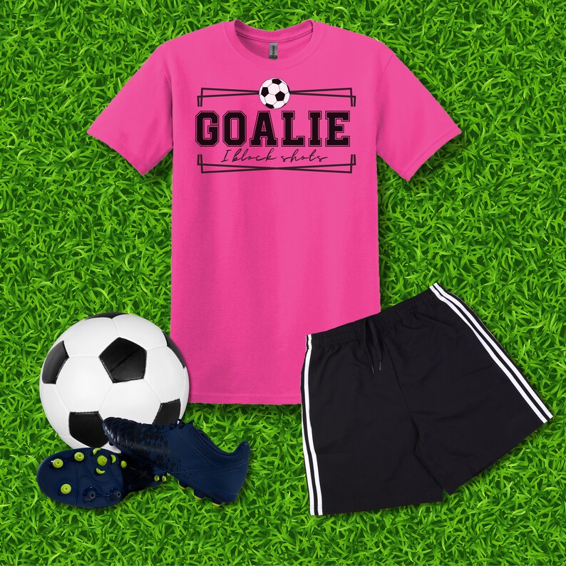 Puede incluir: Camiseta rosa con texto negro que dice "Goalie I block shots" y un gr&aacute;fico de bal&oacute;n de f&uacute;tbol blanco y negro. Un pantal&oacute;n corto deportivo negro con rayas blancas a los lados. Un bal&oacute;n de f&uacute;tbol blanco y negro y un par de tacos de f&uacute;tbol negros y azules.