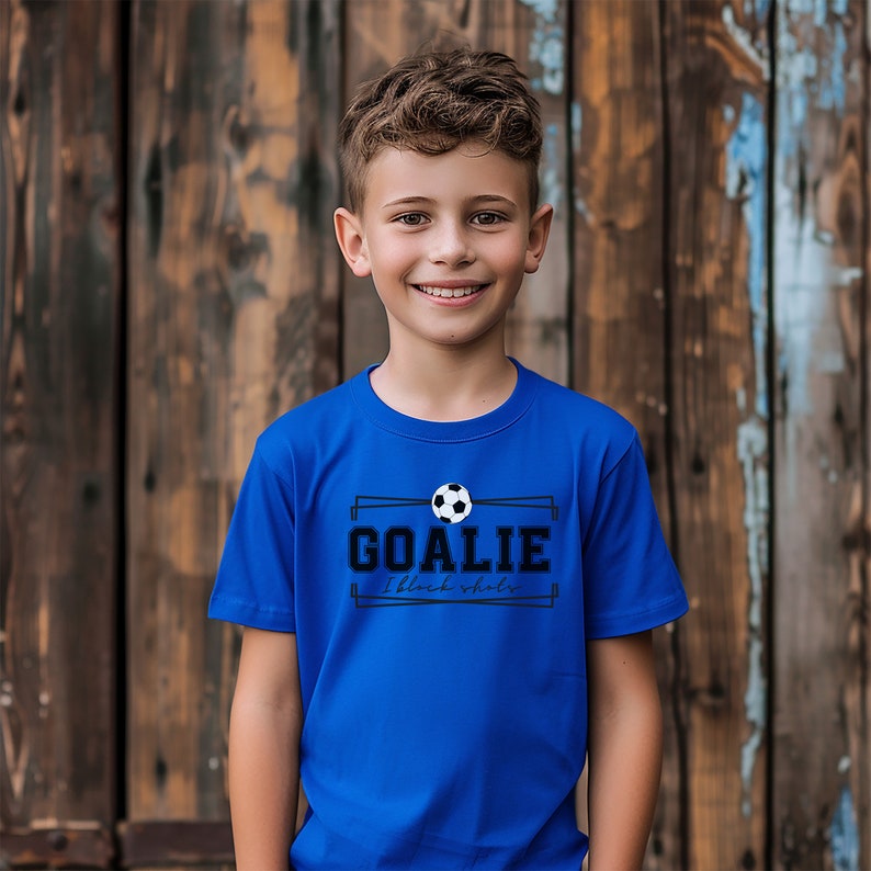 Puede incluir: Un ni&ntilde;o peque&ntilde;o con una camiseta azul real con un gr&aacute;fico de bal&oacute;n de f&uacute;tbol blanco y negro y el texto "GOALIE I block shots".