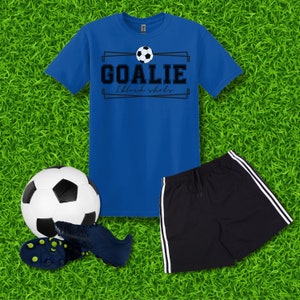 Puede incluir: Una camiseta azul con un gr&aacute;fico de bal&oacute;n de f&uacute;tbol blanco y negro y el texto "Goalie I block shots". Un bal&oacute;n de f&uacute;tbol negro, un par de tacos de f&uacute;tbol negros y pantalones cortos deportivos negros con rayas blancas sobre un fondo de hierba verde.