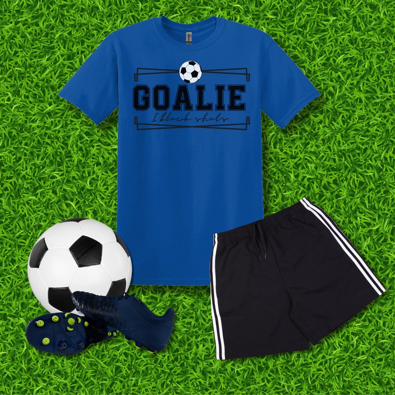 Puede incluir: Una camiseta azul con un gr&aacute;fico de bal&oacute;n de f&uacute;tbol blanco y negro y el texto "Goalie I block shots". Un bal&oacute;n de f&uacute;tbol negro, un par de tacos de f&uacute;tbol negros y pantalones cortos deportivos negros con rayas blancas sobre un fondo de hierba verde.