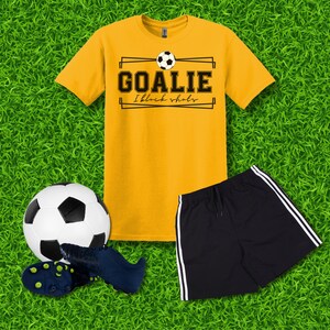 Puede incluir: Una camiseta amarilla con un gr&aacute;fico negro que dice "Goalie I block shots" con un bal&oacute;n de f&uacute;tbol sobre el texto. Un pantal&oacute;n corto deportivo negro con rayas blancas a los lados. Un bal&oacute;n de f&uacute;tbol blanco y negro y un par de botas de f&uacute;tbol negras.