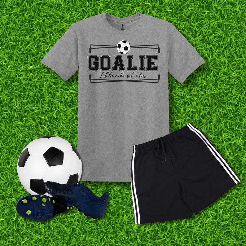 Puede incluir: Una camiseta gris con un gr&aacute;fico de bal&oacute;n de f&uacute;tbol negro y el texto "Goalie I block shots". Un pantal&oacute;n deportivo negro con rayas blancas a los lados. Un bal&oacute;n de f&uacute;tbol blanco y negro y un par de tacos de f&uacute;tbol negros.
