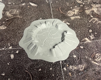 May include: A light gray, circular 3D printed terrain piece featuring a central crater with a cracked, uneven surface texture. The perimeter is surrounded by raised, jagged rock-like formations and concentric topographical contour lines.
