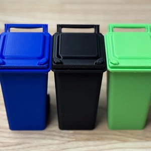 Può includere: Tre bidoni della spazzatura in miniatura, uno blu, uno nero e uno verde. I bidoni della spazzatura sono in plastica e hanno un design semplice.