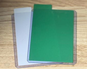 May include: Two trading card sleeves, one with a white card and the other with a green card. The cards are inside clear plastic sleeves. The background is a light brown wooden surface.
