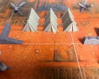 May include: A set of three miniature gray plastic tents arranged in a row, accompanied by gray plastic sandbag wall segments and metal-style anti-tank obstacles, all positioned on a textured orange and brown grid-patterned game board surface.