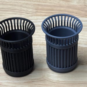 Può includere: Due piccoli bidoni della spazzatura cilindrici. Uno è nero, l'altro è grigio scuro. Entrambi hanno un design a doghe attorno al corpo e un bordo arrotondato con barre verticali. I bidoni sono su una superficie di legno chiaro.