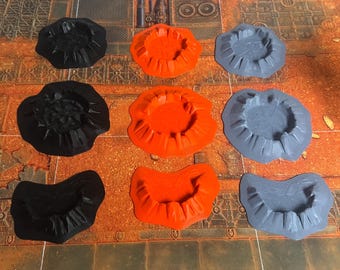 May include: A collection of nine crater-shaped terrain pieces arranged in a three-by-three grid. The items are colored black, bright orange, and light gray, featuring raised, jagged edges and textured, cracked surfaces resembling impact sites.