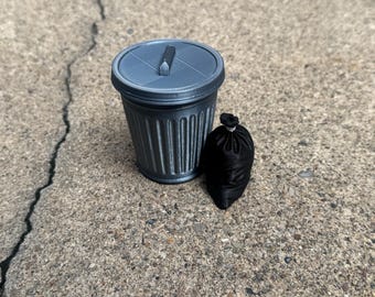 May include: A miniature grey plastic trash can with a removable lid and a small black fabric drawstring bag positioned next to it. The trash can features vertical ridges along the sides and a central handle on the circular lid, while the bag is tied.