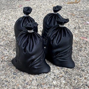 May include: Four small, black, sack-shaped objects with a textured surface. Each sack is gathered at the top and secured with a small, white, cylindrical ring, creating a distinct bunched appearance at the neck of each individual container.