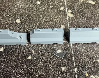 May include: Five miniature grey plastic jersey barriers arranged in a horizontal row. Each barrier features a sloped profile with a flat top, a wide base, and a small loop handle on the upper edge, designed for tabletop gaming terrain setups.