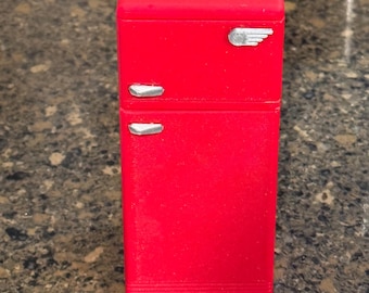 May include: A bright red miniature refrigerator with silver-colored accents. The small appliance has a rectangular shape with a top and bottom section, and a silver emblem on the top door. The refrigerator is a retro design.
