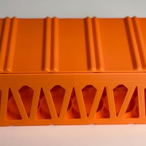 May include: An orange rectangular storage box with a ridged top and geometric cutouts along the sides. The box is made of a solid, opaque material and sits on a white surface.