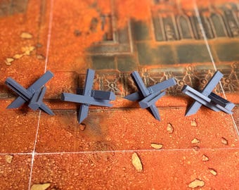 May include: Four grey, three-dimensional, cross-shaped anti-tank obstacles, known as Czech hedgehogs, arranged in a horizontal row. Each piece features three intersecting beams with sharp, pointed ends, designed for tabletop gaming terrain.
