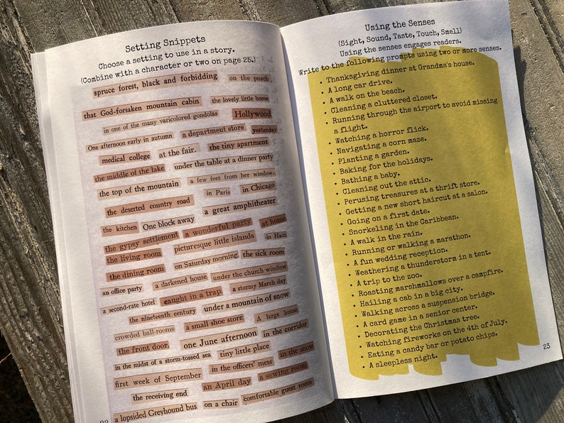 May include: An open book with two pages of writing exercises. The left page, titled Setting Snippets, contains numerous small rectangular paper strips with printed phrases. The right page, titled Using the Senses, features a yellow box with a list of prompts.