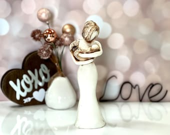 May include: White ceramic figurine of a mother holding a baby. The figurine is standing on a white surface with a wooden heart that says "xoxo" and a metal sign that says "Love".