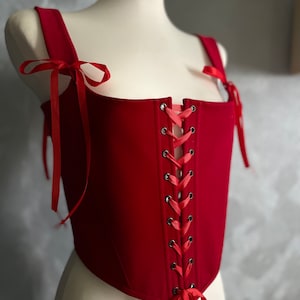 May include: A red corset with a lace-up back and ribbon ties. The corset is made of a thick fabric and has a fitted silhouette.