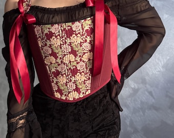 May include: A burgundy corset with a floral pattern in cream and green, accented with red ribbons. The corset has a structured bodice and is paired with a black off-the-shoulder top and black lace shorts. The overall style is gothic and romantic.
