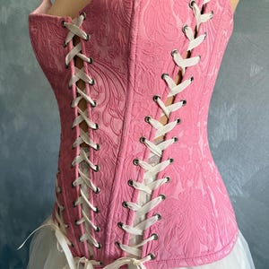 May include: A pink corset with a floral pattern and white lacing. The corset is paired with a white tulle skirt, creating a romantic and feminine aesthetic. The corset features metal eyelets and white ribbon ties.