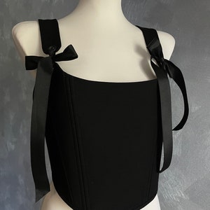 May include: A black corset top with a square neckline and adjustable straps that tie into bows. The straps are made of black satin ribbon.