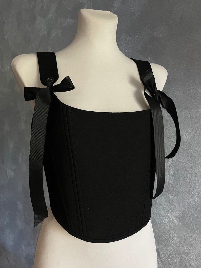 May include: A black corset top with a square neckline and adjustable straps that tie into bows. The straps are made of black satin ribbon.