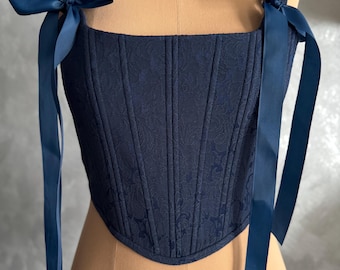 May include: Navy blue corset with a floral pattern. The corset features a square neckline, structured boning, and shoulder straps tied with navy blue ribbons. The garment is displayed on a mannequin.