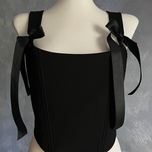 May include: A black corset top with a square neckline and two black ribbon straps that tie into bows at the shoulders.