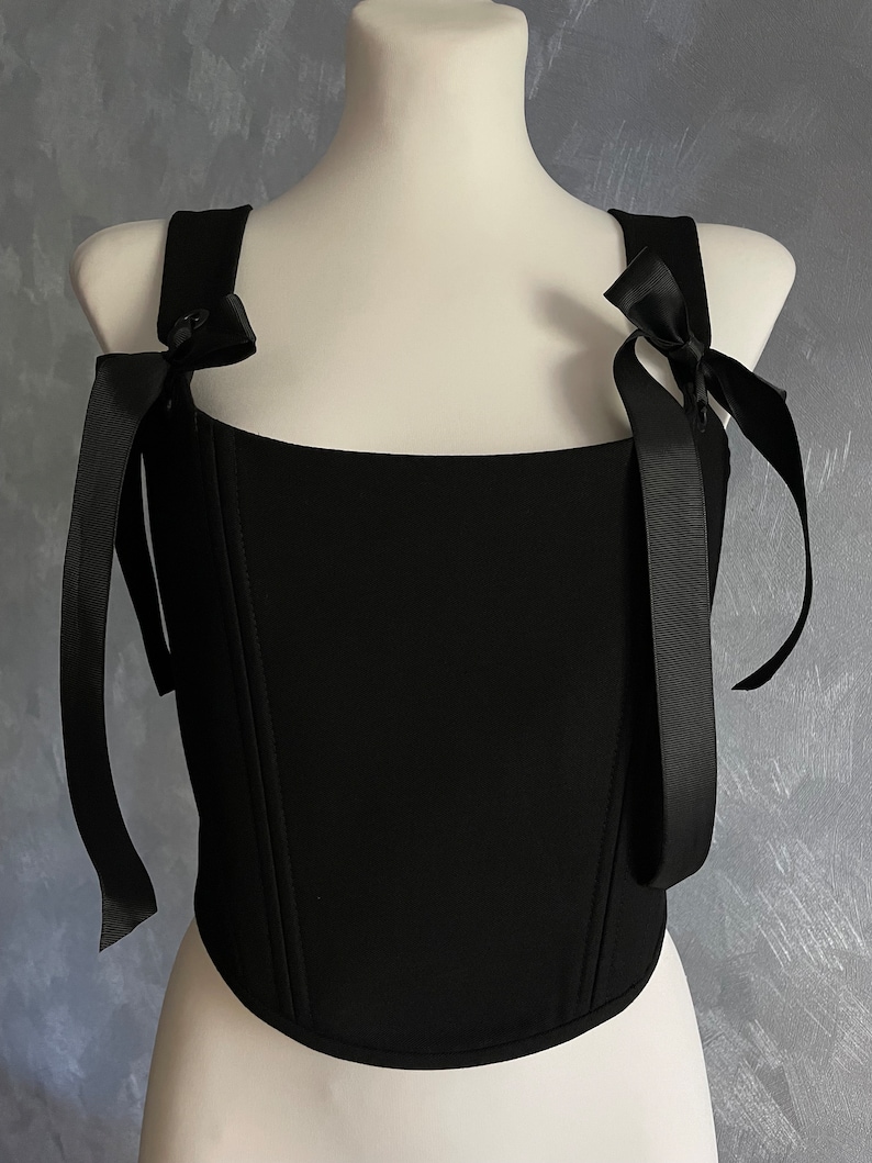 May include: A black corset top with a square neckline and two black ribbon straps that tie into bows at the shoulders.