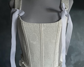 May include: White corset with a floral pattern and white ribbon straps. The corset has a ruffled bottom.