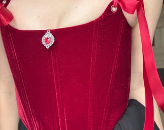 May include: A deep red velvet corset with ribbon ties and a decorative jewel pendant. The corset features a sweetheart neckline and is paired with a matching choker necklace. The straps are adorned with bows.