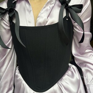 May include: Black corset with attached shoulder straps tied with black bows. The corset is worn over a light purple button-down shirt. The straps are attached to the corset with metal eyelets.