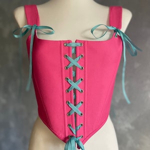 May include: A pink corset with a blue ribbon lacing up the front. The corset has a pointed bottom and is made of a smooth fabric.