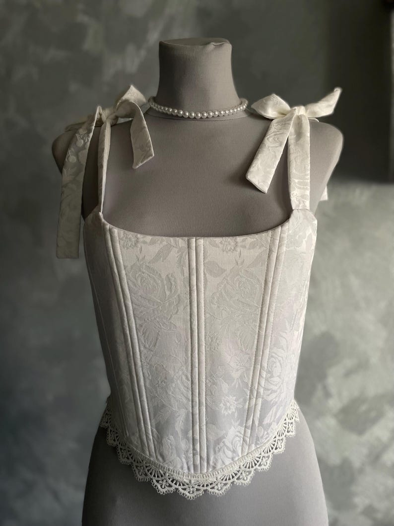 Op de afbeelding: Witte corset met een bloemenpatroon en kantversiering. De corset heeft verstelbare bandjes die aan de schouders worden vastgebonden.