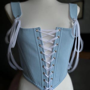 May include: A light blue corset with white ribbon straps and white laces. The corset is laced up the front with metal eyelets.
