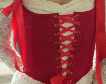 May include: A red corset featuring a subtle tonal damask pattern, a front lace-up closure with metal grommets, and red satin ribbon ties at the shoulders and bottom. The corset is worn over a white long-sleeved top with a textured lace fabric.