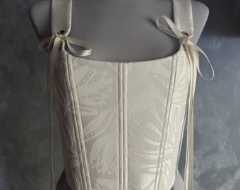 May include: Ivory-colored corset with a floral pattern, displayed on a mannequin. The corset features structured boning, shoulder straps, and delicate ribbon ties. A white lace choker is around the mannequin's neck. The overall design is elegant and vintage-inspired.