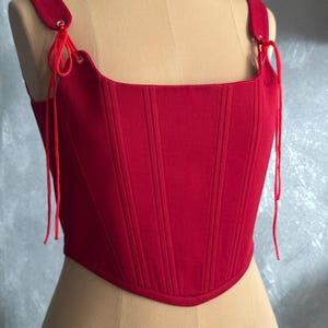 May include: A bright red corset-style top featuring a square neckline and vertical stitched paneling. The garment includes shoulder straps with silver-toned metal eyelets and matching red cord ties that hang down on both sides of the bodice.