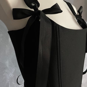May include: Black corset top with adjustable straps and a bow detail. The straps have a metal ring and buckle closure.