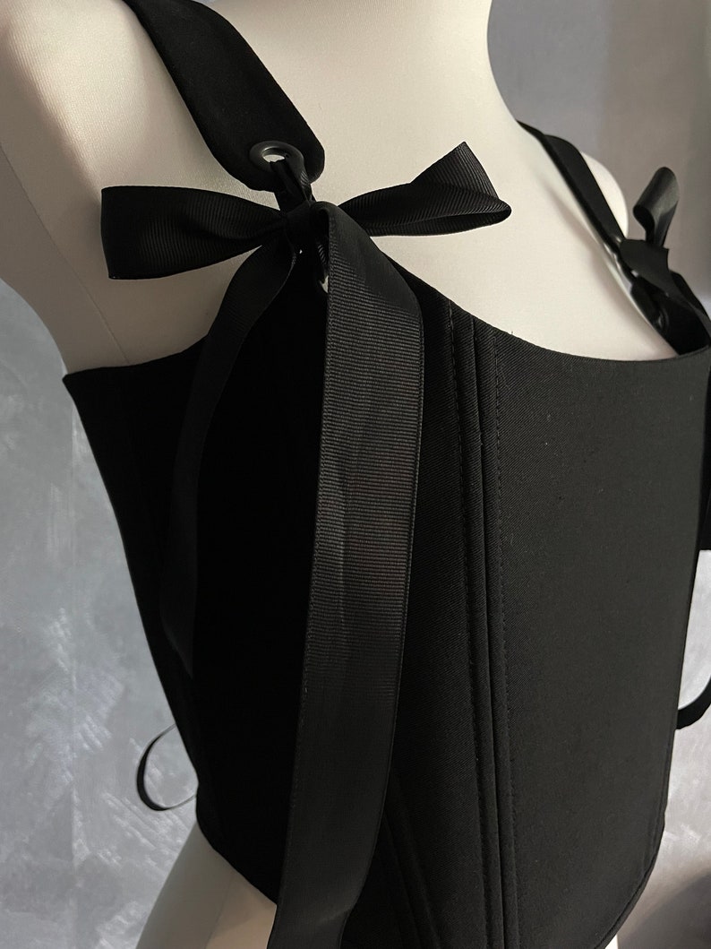 May include: Black corset top with adjustable straps and a bow detail. The straps have a metal ring and buckle closure.